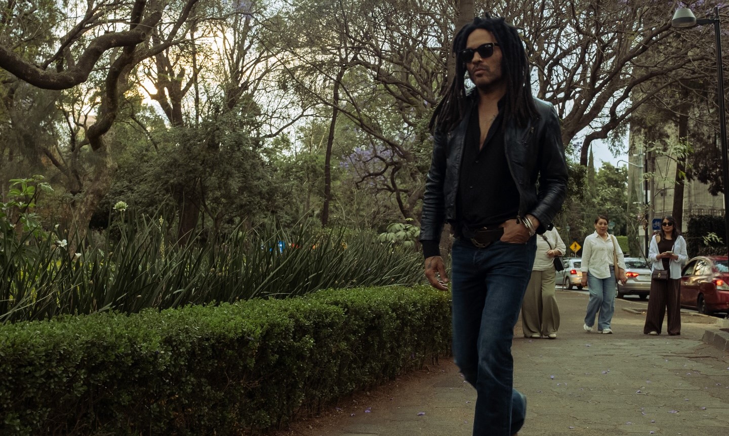 Retrato de cuerpo entero del músico Lenny Kravitz caminando con seguridad por una acera adoquinada en la Ciudad de México. Viste una chaqueta de cuero negra abierta, camisa negra, pantalones vaqueros de campana y botas de gamuza. Lleva gafas de sol y su peinado rastas es visible. El fondo está dominado por árboles de jacarandá con follaje verde y flores de color lila suave contra un cielo luminoso. Varias personas caminan detrás de él en la distancia.