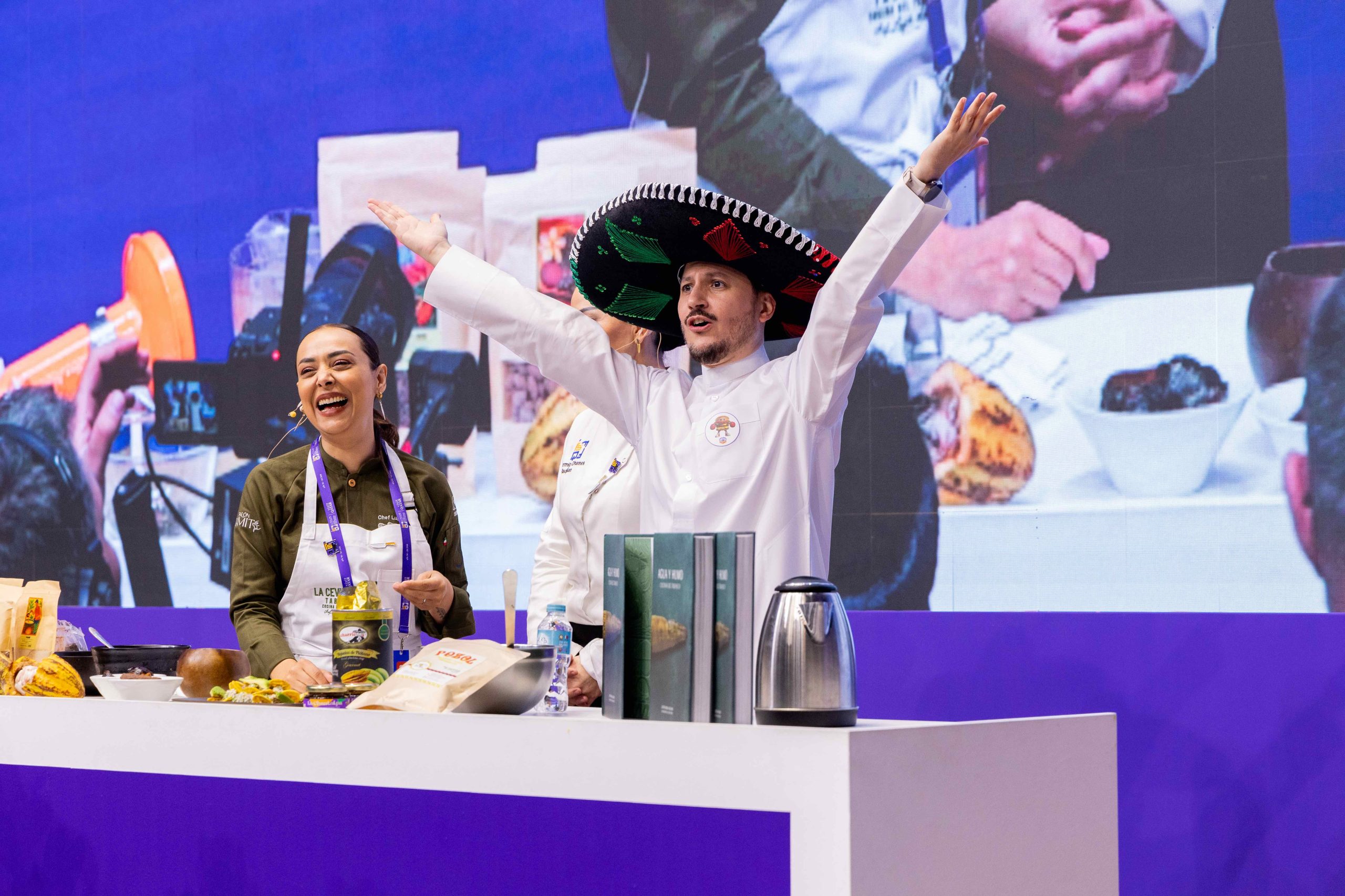 La Chef Lupita Vidal Sonríe Durante Su Clase Magistral En El Qatar International Food Festival 2026, Acompañada Por Un Chef Con Sombrero Charro Y Ejemplares De Su Libro &Quot;Agua Y Humo&Quot; Frente A Una Audiencia Internacional En Doha.