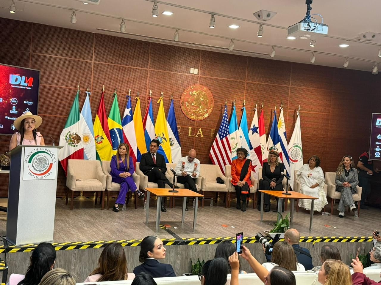 El senado de la república se viste de gala: celebran el liderazgo de las 100 mujeres de américa latina 2026 la revista del sureste mx Fotografía Del Escenario Principal En El Auditorio Octavio Paz Del Senado De La República De México. En El Centro, Sobre Una Pared De Madera, Destaca El Emblema Nacional Mexicano Junto A Una Fila De Banderas De América Latina, Incluyendo México, Chile, Estados Unidos Y Otros. Al Podio De La Izquierda, La Alcaldesa De Uruapan, Grecia Quiroz, Con Un Sombrero, Ofrece Un Discurso.
