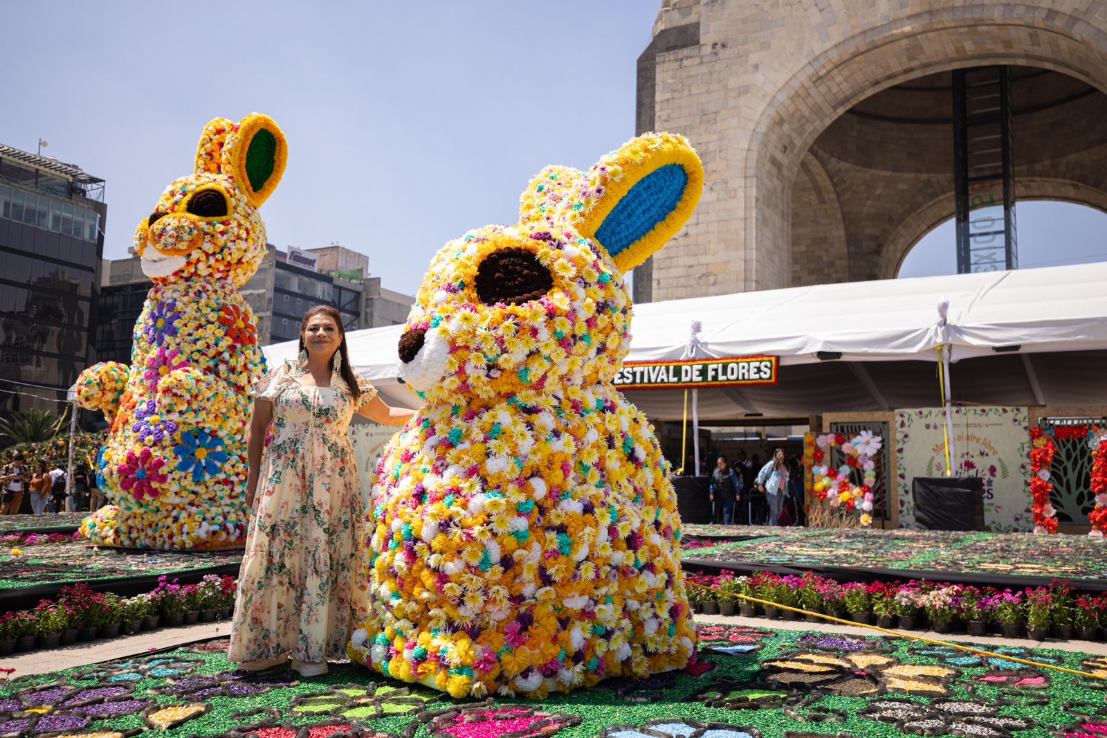 : Clara Brugada, Jefa de Gobierno de la Ciudad de México, sonríe de pie entre dos enormes esculturas de conejos hechas de flores en el Monumento a la Revolución. Viste un vestido largo floreado. Los conejos, coloridos con flores amarillas, moradas y rosas, representan teporingos, una especie endémica, y son parte del Primer Festival de las Flores de Primavera 2026. Detrás de ella, se ve el arco del Monumento y una carpa blanca con el letrero "FESTIVAL DE FLORES". El suelo tiene alfombras florales. La imagen destaca el evento cultural y turístico.