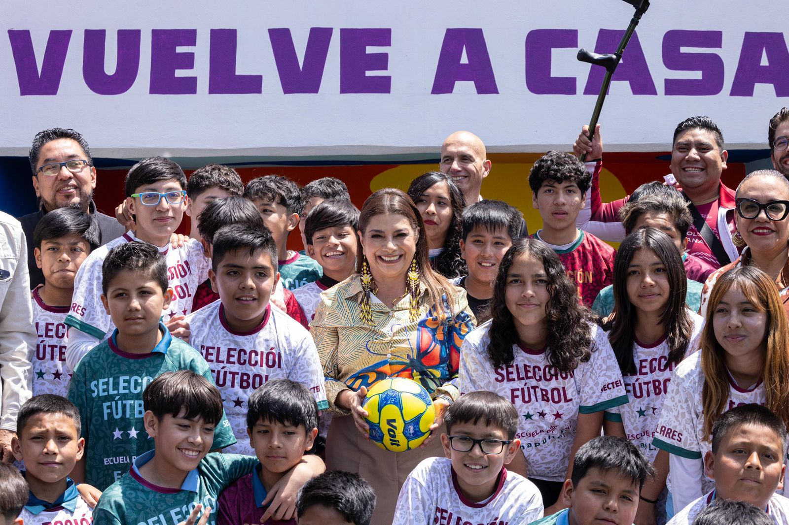 La Jefa de Gobierno de la CDMX, Clara Brugada, sonríe rodeada de niños y jóvenes futbolistas de Iztapalapa. Brugada sostiene un balón de fútbol amarillo frente a un muro con la frase "VUELVE A CASA" en letras moradas.