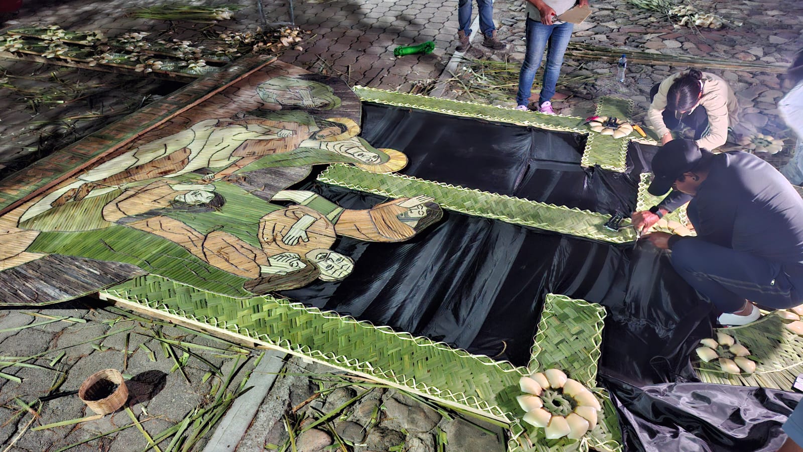 Fotografía En Plano Picado (Desde Arriba) Que Captura El Intrincado Proceso De Elaboración De Un Arco Monumental De Cucharilla En Un Suelo De Adoquín. En Primer Plano, Una Gran Obra De Arte Sacro Está Siendo Tejida Y Ensamblada.