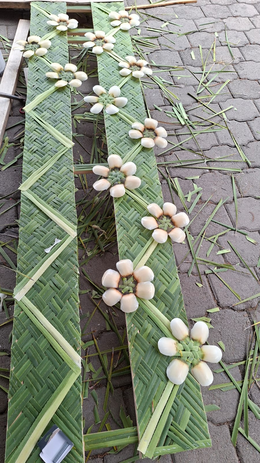 Fotografía En Primer Plano De Un Panel De Madera Cubierto Con Un Intrincado Tejido De Hojas De Palma Verde Claro, En Proceso De Elaboración De Un Arco Monumental De Cucharilla. El Tejido Presenta Patrones Geométricos Intrincados En Diagonal. A Lo Largo Del Centro Del Panel, Una Hilera De &Quot;Flores De Cucharilla&Quot; Artesanales Está Fijada A La Superficie.