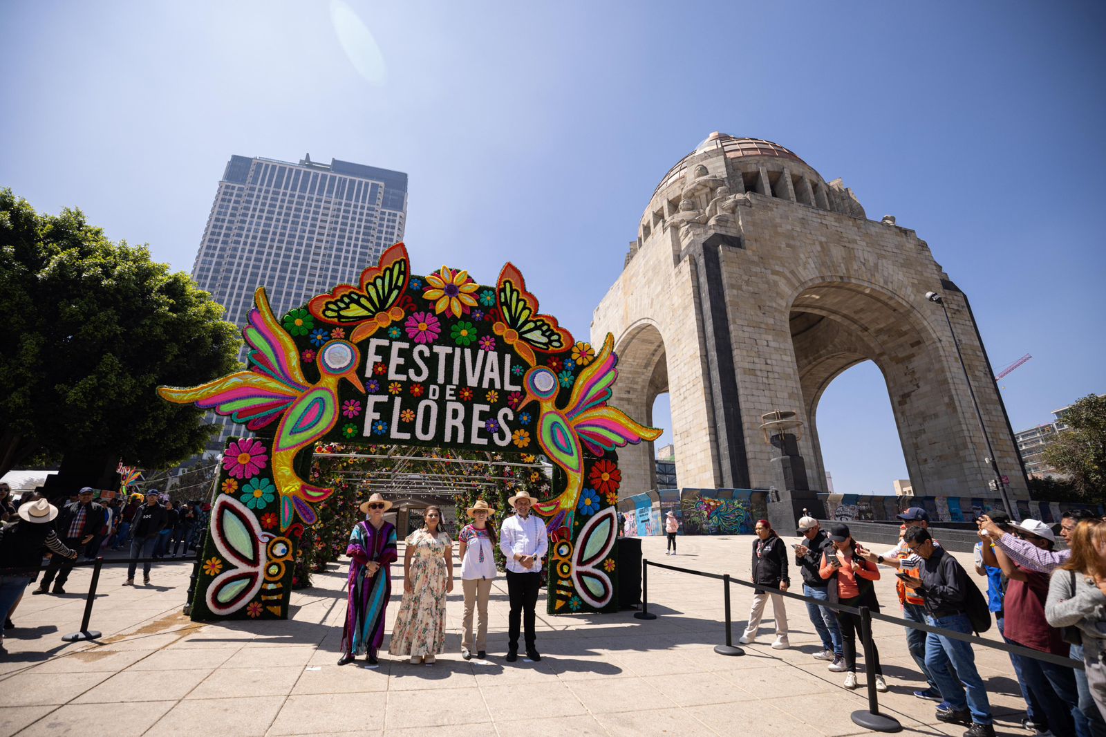 La Jefa De Gobierno De La Cdmx, Clara Brugada, Junto A Funcionarios, Posan Frente A Un Monumental Arco De Flores Con La Inscripción &Quot;Festival De Flores&Quot; En La Explanada Del Monumento A La Revolución. El Arco Está Decorado Con Figuras De Mariposas, Colibríes Y Flores Multicolores. De Fondo Se Aprecia El Arco Del Monumento Y Edificios Del Centro De La Ciudad Bajo Un Cielo Despejado.
