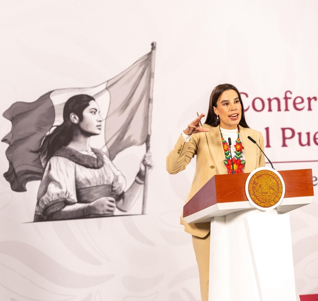 Josefina Rodríguez Zamora, Titular De La Secretaría De Turismo De México (Sectur), De Pie Detrás De Un Podio Con El Escudo Nacional. Viste Un Traje Sastre Color Arena Y Un Collar De Flores Artesanales En Tonos Rojo Y Verde