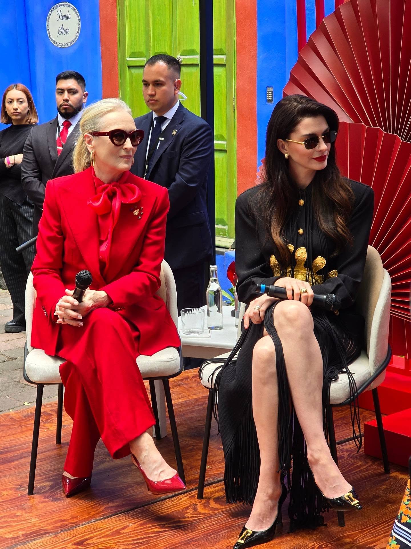 Fotografía De La Conferencia De Prensa En La Casa Azul De La Cdmx, Con Meryl Streep Y Anne Hathaway Sentadas En El Centro. Meryl Streep Luce Un Traje Sastre Rojo Brillante Con Una Lazada Al Cuello Y Lentes Oscuros, Mientras Que Anne Hathaway Viste Un Elegante Diseño Negro Con Detalles Dorados Y Flecos Largos.