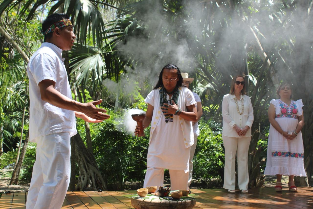 "Cuatro personas participan en una ceremonia tradicional maya al aire libre, rodeados de densa vegetación tropical. Un hombre maya en primer plano, con vestimenta blanca y banda de cabeza colorida, mira hacia la izquierda. En el centro, otro hombre maya vestido de blanco y con collar verde sostiene un cuenco de cerámica humeante de incienso de copal sobre un altar de piedra con jícaras y ofrendas. Detrás de ellos, una turista con camisa blanca y pantalones blancos, y otra mujer con un vestido bordado tradicional maya, observan la ceremonia con respeto. El fondo es un jardín de palmeras y árboles frondosos."