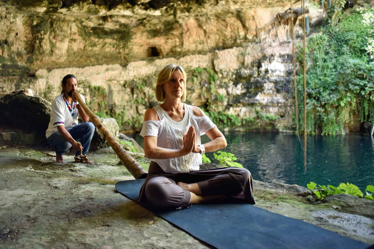 Una turista realiza una sesión de meditación y yoga en pose de loto, con las manos en oración, sobre un tapete negro a la orilla de un cenote de agua azul cristalina en Yucatán. Detrás de ella, un facilitador local con vestimenta tradicional mayas se encuentra en cuclillas tocando un didgeridoo (instrumento de viento largo de madera). Esta experiencia de bienestar integral y conexión ancestral es parte de las actividades ofrecidas durante el Maya Wellness Weekend en locaciones sostenibles y con identidad local en México."
