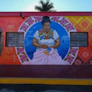 Caminos ancestrales: el nuevo corredor de arte y cultura maya en felipe carrillo puerto la revista del sureste mx Fotografía De Frente Que Captura Un Mural Artístico Vibrante Pintado En La Fachada De Un Edificio De Una Sola Planta En Felipe Carrillo Puerto. El Mural Está Dividido En Tres Secciones Principales. La Sección Central Muestra Un Gran Retrato Circular De Una Mujer Maya De Mediana Edad Con Un Peinado Recogido, Vistiendo Un Huipil Tradicional Blanco Bordado Con Flores De Colores.
