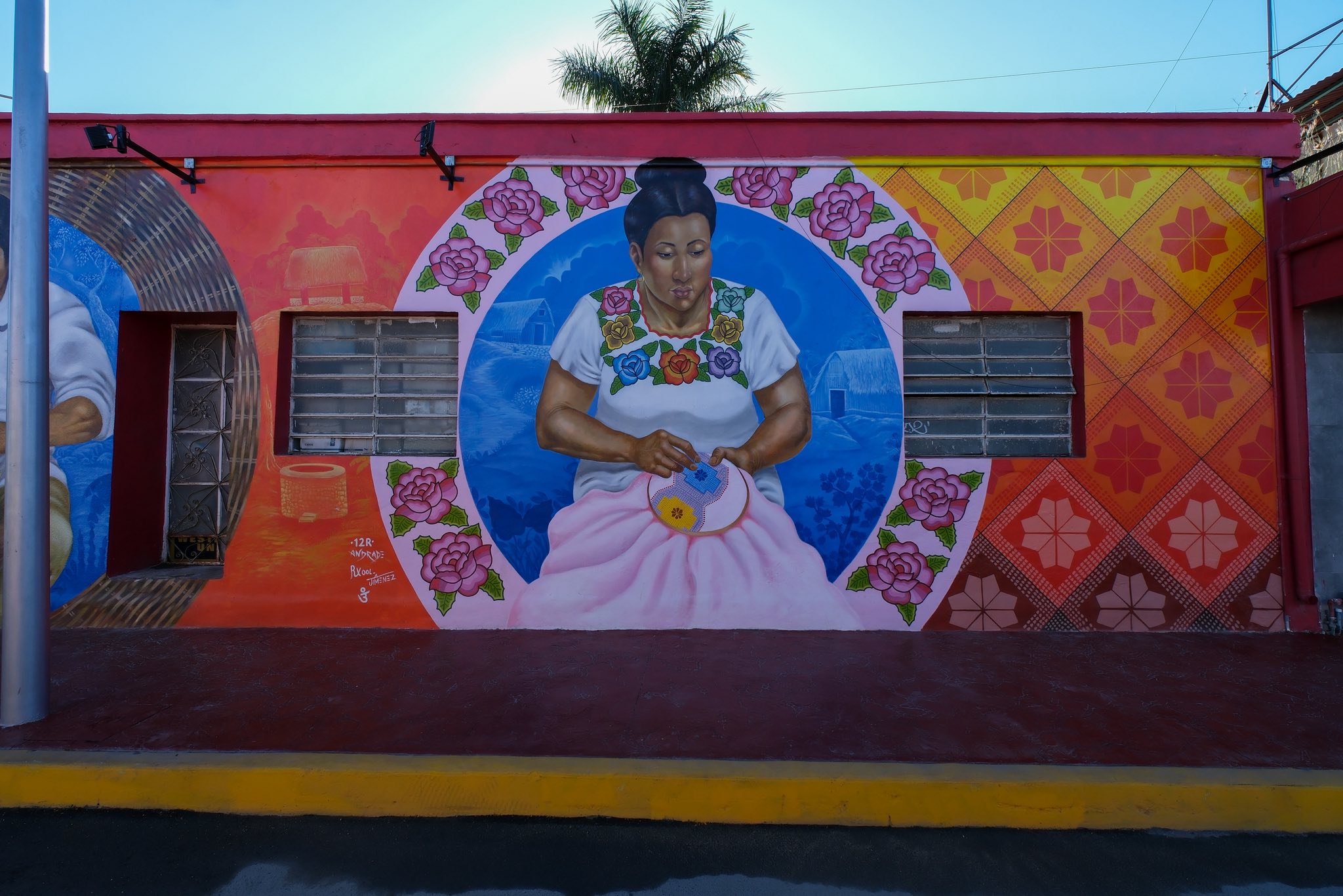 Fotografía De Frente Que Captura Un Mural Artístico Vibrante Pintado En La Fachada De Un Edificio De Una Sola Planta En Felipe Carrillo Puerto. El Mural Está Dividido En Tres Secciones Principales. La Sección Central Muestra Un Gran Retrato Circular De Una Mujer Maya De Mediana Edad Con Un Peinado Recogido, Vistiendo Un Huipil Tradicional Blanco Bordado Con Flores De Colores.