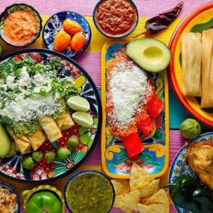 Fotografía Cenital (Desde Arriba) De Un Banquete De Comida Mexicana Servido Sobre Una Mesa De Madera Con Franjas De Colores Vibrantes (Rosa, Amarillo, Azul). Se Aprecian Diversos Platillos Tradicionales En Vajilla De Talavera Y Barro: Un Plato Grande Con Enchiladas Verdes Cubiertas De Queso Y Crema, Tamales En Sus Hojas De Maíz, Chiles Rellenos, Totopos, Guacamole En Un Molcajete De Piedra, Y Diversas Salsas Roja Y Verde.