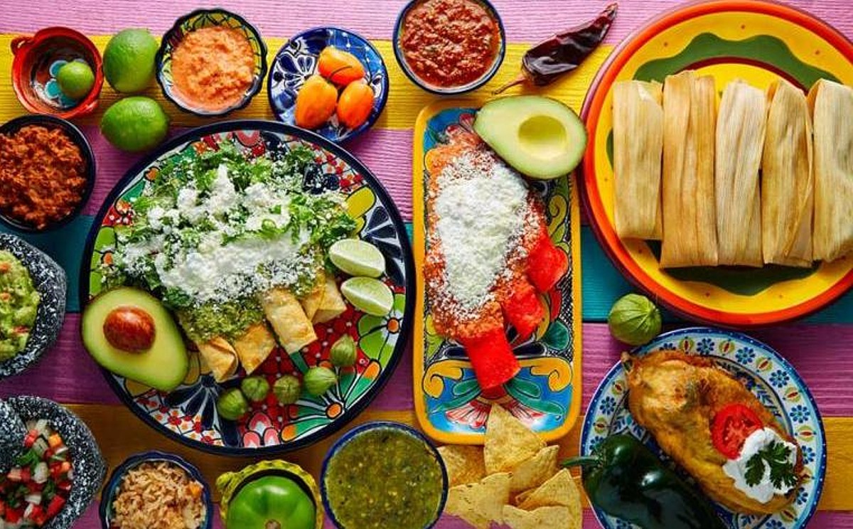 Fotografía Cenital (Desde Arriba) De Un Banquete De Comida Mexicana Servido Sobre Una Mesa De Madera Con Franjas De Colores Vibrantes (Rosa, Amarillo, Azul). Se Aprecian Diversos Platillos Tradicionales En Vajilla De Talavera Y Barro: Un Plato Grande Con Enchiladas Verdes Cubiertas De Queso Y Crema, Tamales En Sus Hojas De Maíz, Chiles Rellenos, Totopos, Guacamole En Un Molcajete De Piedra, Y Diversas Salsas Roja Y Verde.