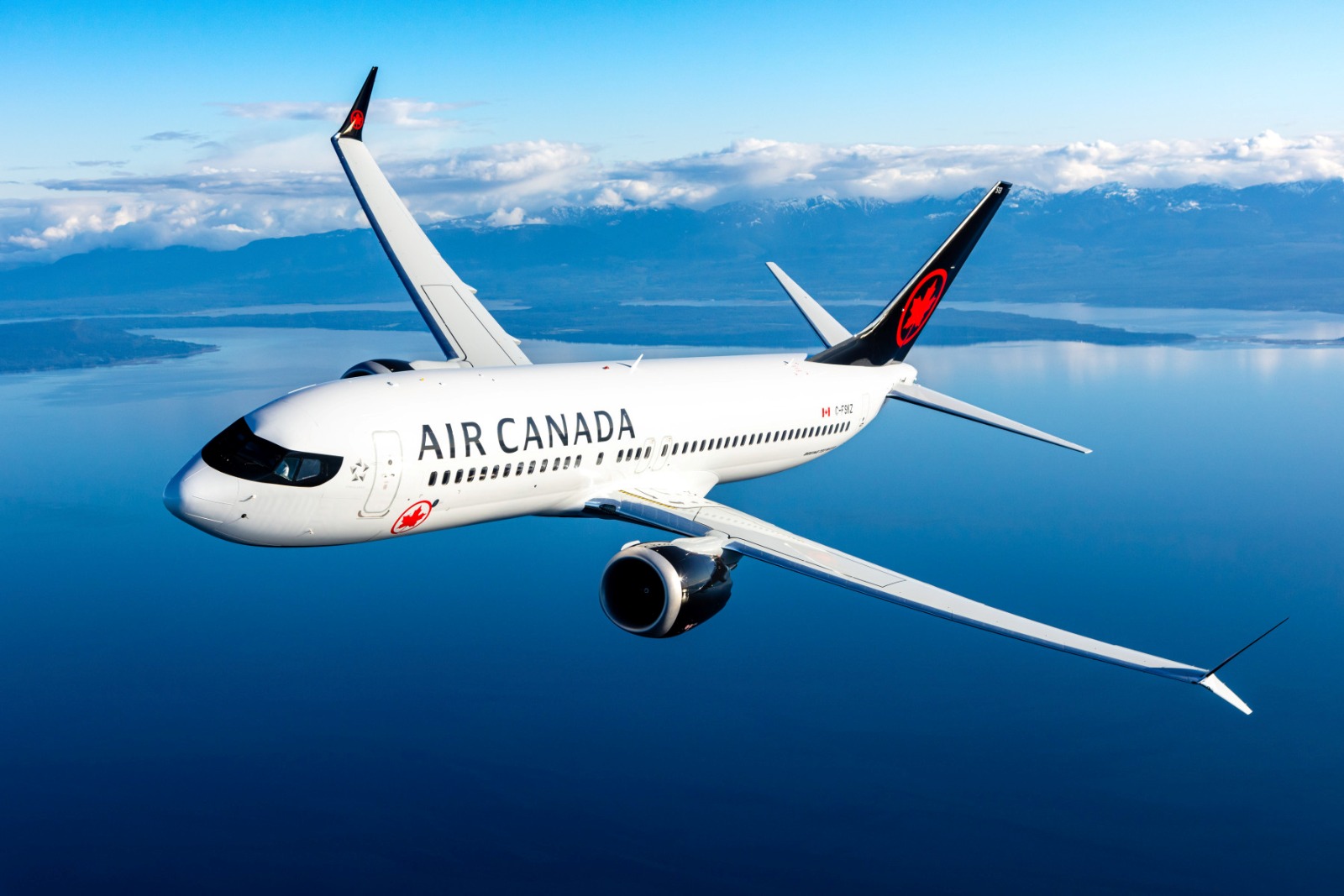 Una Fotografía Aérea De Plano Medio Que Captura Un Avión De Pasajeros Boeing 737 Max De Air Canada En Pleno Vuelo. El Avión, Pintado De Blanco Con La Palabra &Quot;Air Canada&Quot; En Letras Negras Mayúsculas En El Fuselaje Y El Logotipo De La Hoja De Arce Roja En La Cola Negra, Vuela Sobre Un Vasto Cuerpo De Agua De Color Azul Profundo Que Refleja El Cielo.