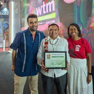 Co’ox mayab: el rugido del turismo comunitario de yucatán resuena en el escenario mundial de brasil la revista del sureste mx Una Fotografía De Tres Personas De Pie En El Stand De Wtm Latin America. Al Centro, Ismael Arellano Ciau, Secretario De Co’ox Mayab, Viste Una Guayabera Blanca Con Bordados Tradicionales Y Sostiene El Diploma Enmarcado Del Premio De Bronce.
