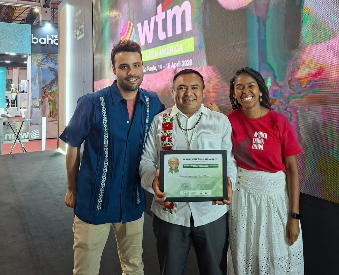 Co’ox mayab: el rugido del turismo comunitario de yucatán resuena en el escenario mundial de brasil la revista del sureste mx Una Fotografía De Tres Personas De Pie En El Stand De Wtm Latin America. Al Centro, Ismael Arellano Ciau, Secretario De Co’ox Mayab, Viste Una Guayabera Blanca Con Bordados Tradicionales Y Sostiene El Diploma Enmarcado Del Premio De Bronce.