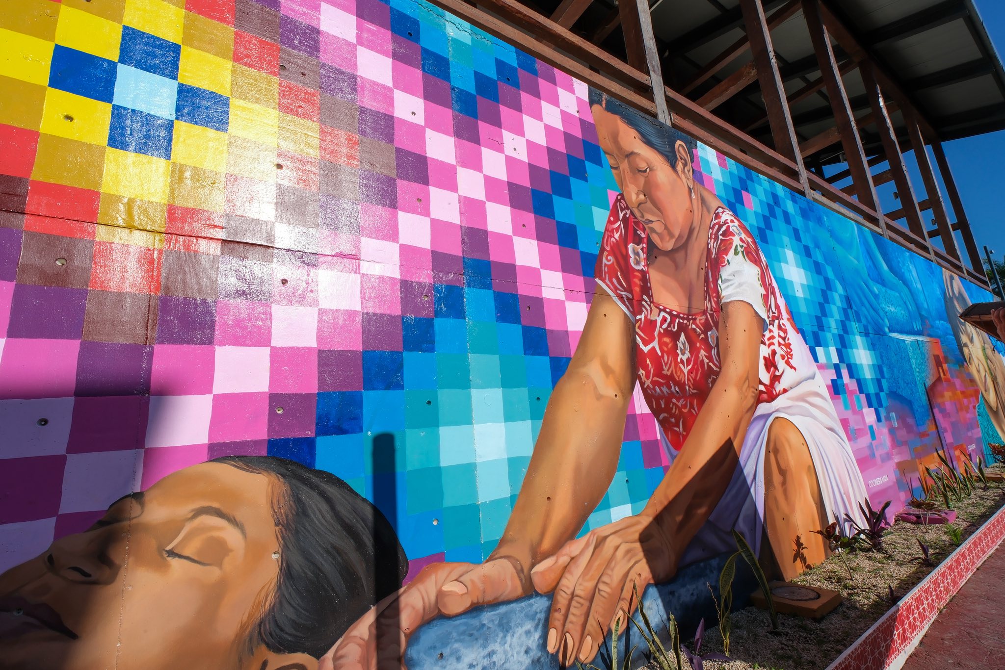 Caminos ancestrales: el nuevo corredor de arte y cultura maya en felipe carrillo puerto la revista del sureste mx Un Mural De Arte Callejero De Plano Medio Capturado Con Un Ángulo Ligeramente Contrapicado En Felipe Carrillo Puerto. A La Izquierda, El Rostro De Una Joven Mujer Maya En Reposo, Con Los Ojos Cerrados, Ocupa El Primer Plano. El Resto Del Mural Está Dominado Por La Figura De Una Partera Maya Anciana, Arrodillada Y Concentrada, Vestida Con Un Huipil Tradicional Rojo Bordado.