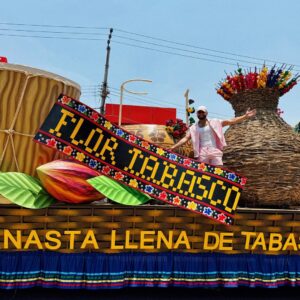 Eli zamora en el desfile de carros alegóricos de la feria tabasco 2026 la revista del sureste mx Fotografía Del Cantante Y Creador De Contenido Eli Zamora, Vistiendo Un Conjunto Rosa Claro Y Gorra Blanca, Posando Con Los Brazos Extendidos Sobre Un Monumental Carro Alegórico Durante El Desfile De La Feria Tabasco 2026. La Estructura Artesanal Muestra Elementos Icónicos Como Un Tambor Típico, Un Grano De Cacao Gigante, Una Flor De Color Intenso Y Una Enorme Canasta De Mimbre Con Flores. En El Centro Destaca Un Letrero Con La Leyenda &Amp;Quot;Flor Tabasco&Amp;Quot;.