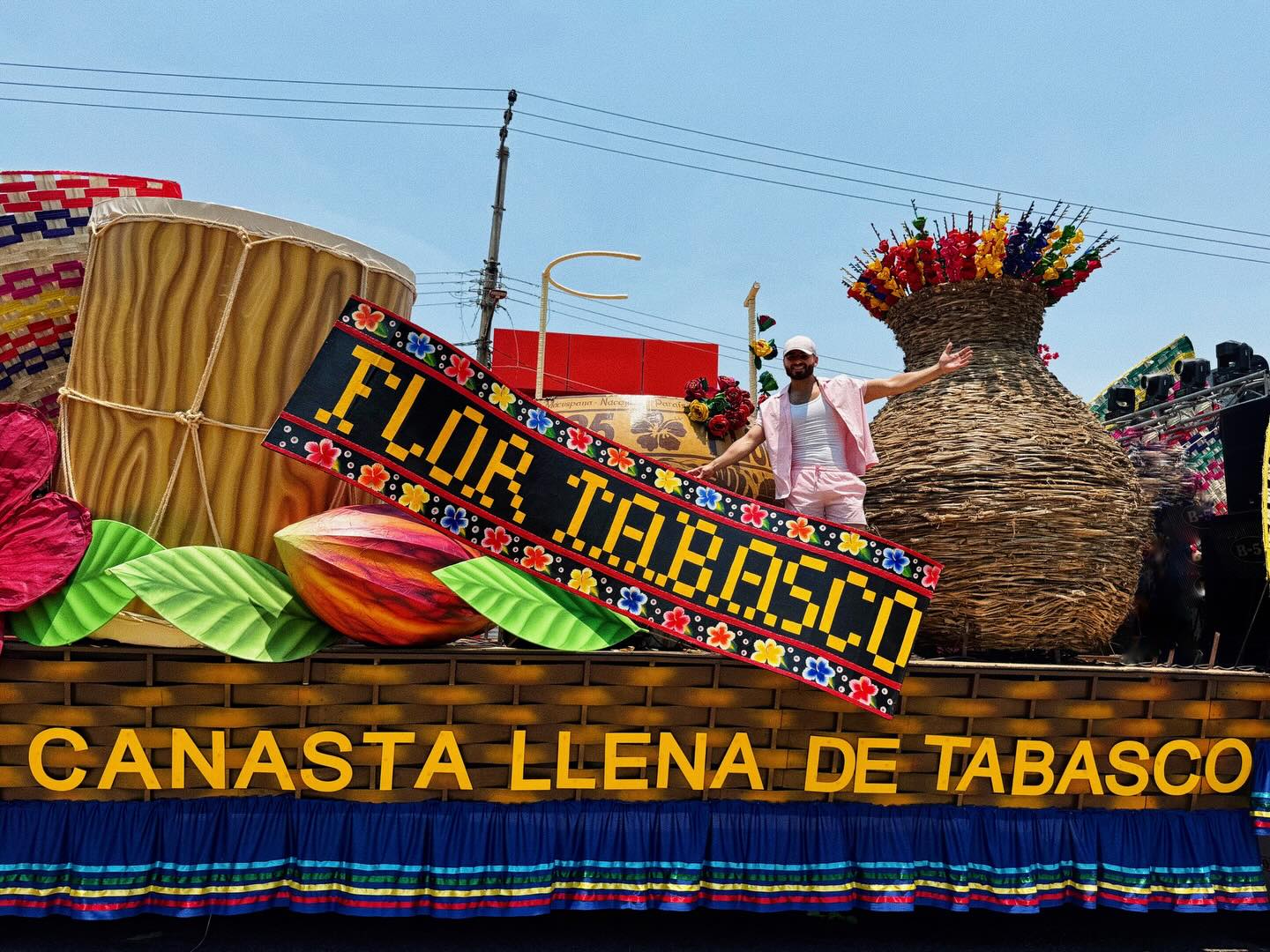 Fotografía Del Cantante Y Creador De Contenido Eli Zamora, Vistiendo Un Conjunto Rosa Claro Y Gorra Blanca, Posando Con Los Brazos Extendidos Sobre Un Monumental Carro Alegórico Durante El Desfile De La Feria Tabasco 2026. La Estructura Artesanal Muestra Elementos Icónicos Como Un Tambor Típico, Un Grano De Cacao Gigante, Una Flor De Color Intenso Y Una Enorme Canasta De Mimbre Con Flores. En El Centro Destaca Un Letrero Con La Leyenda &Quot;Flor Tabasco&Quot;.