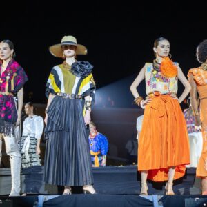 Tres Modelos Desfilan En Una Pasarela Nocturna Al Aire Libre Durante El Evento 'Guerrero, El Origen'. Visten Diseños Contemporáneos Con Influencias Tradicionales: Una Túnica Negra Y Fucsia Con Botas Blancas, Un Conjunto De Rayas Con Sombrero De Ala Ancha, Y Un Atuendo Con Falda Naranja Vibrante. Al Fondo Se Observa El Público Y La Iluminación Del Museo Fuerte De San Diego
