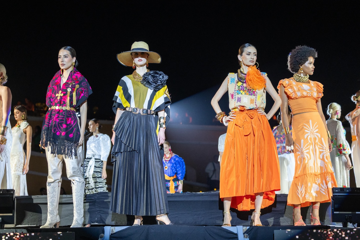 Tres Modelos Desfilan En Una Pasarela Nocturna Al Aire Libre Durante El Evento 'Guerrero, El Origen'. Visten Diseños Contemporáneos Con Influencias Tradicionales: Una Túnica Negra Y Fucsia Con Botas Blancas, Un Conjunto De Rayas Con Sombrero De Ala Ancha, Y Un Atuendo Con Falda Naranja Vibrante. Al Fondo Se Observa El Público Y La Iluminación Del Museo Fuerte De San Diego