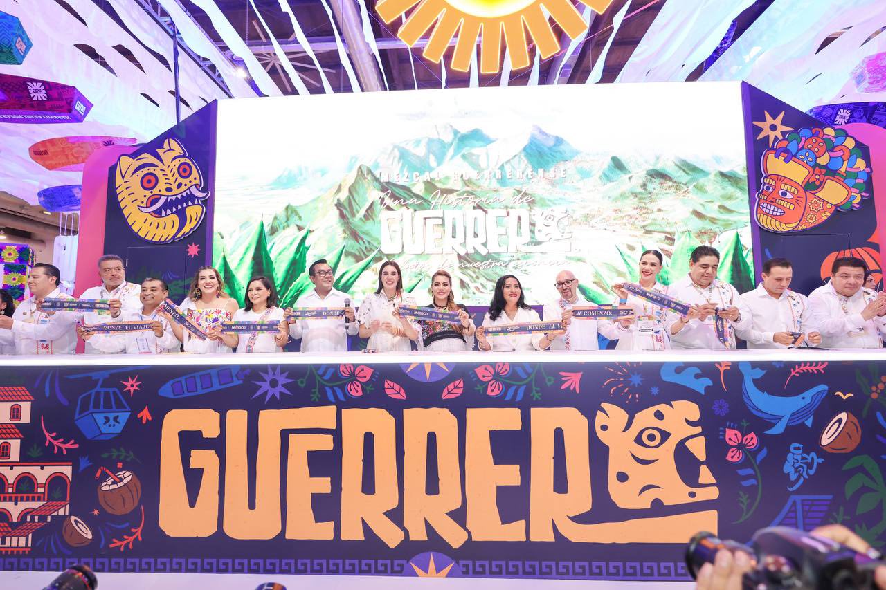 Una Toma Amplia Y Colorida Del Stand De Guerrero En El Tianguis Turístico. Al Frente, Trece Personas —Ocho Hombres Y Cinco Mujeres— Están Alineadas De Pie Detrás De Una Larga Mesa, Sosteniendo Listones Ceremoniales Conmemorativos Mientras Posan Para Una Fotografía Oficial.