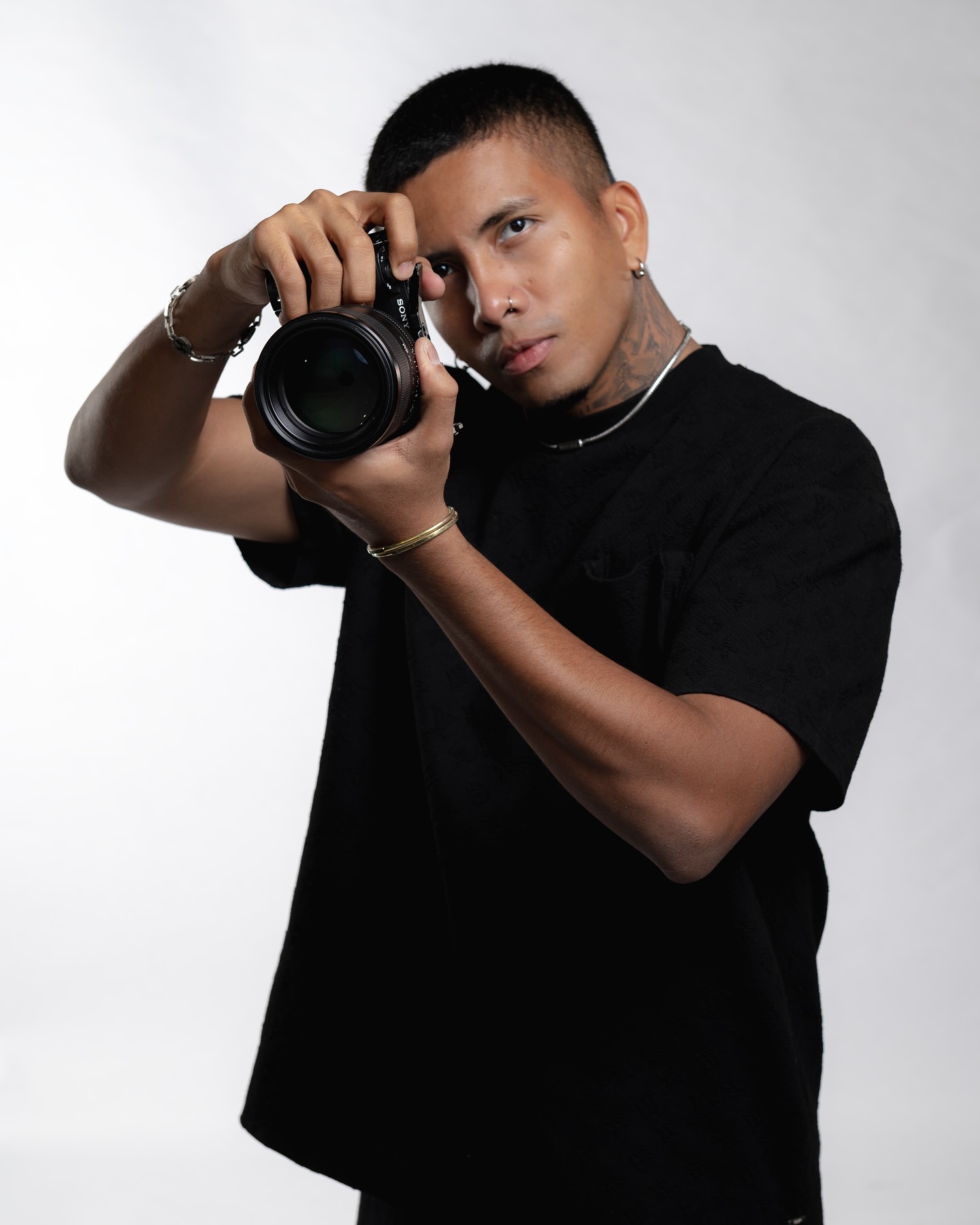 Retrato De Estudio Del Fotógrafo Juan Barcasnegras Sobre Un Fondo Blanco. El Creativo Panameño Viste Una Playera Negra, Luce Tatuajes En El Cuello Y Accesorios Como Cadenas Y Pulseras.
