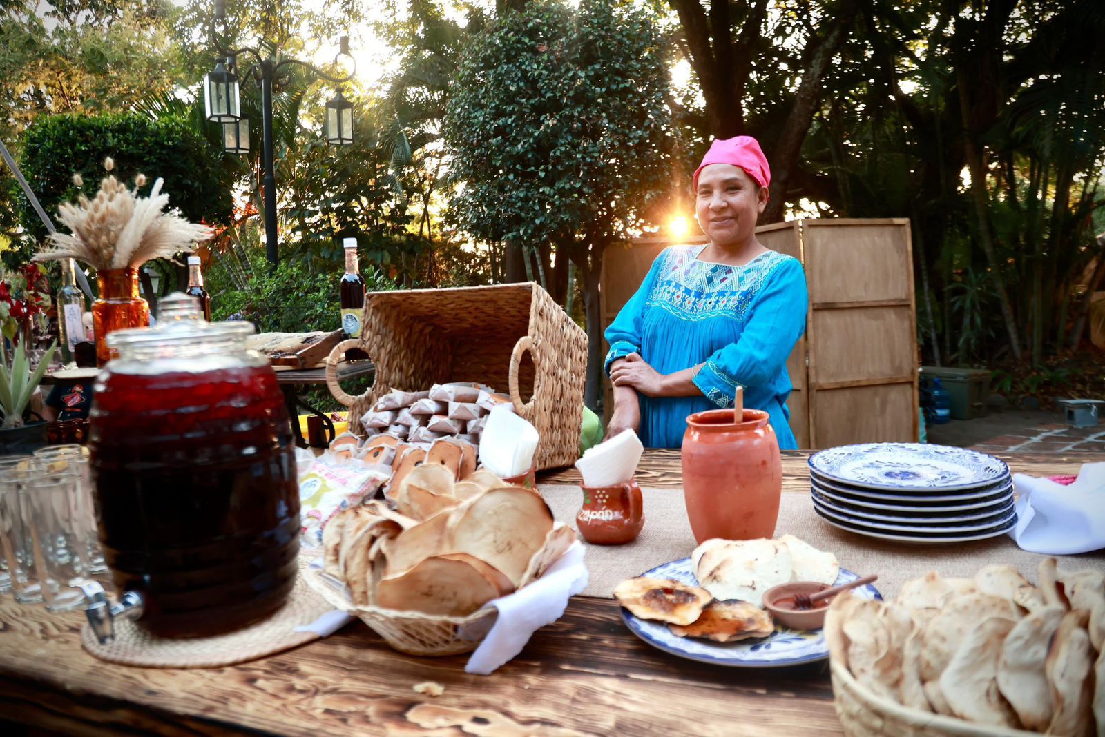 Una Fotografía Cálida Al Aire Libre Que Muestra A Una Cocineras Tradicional Mexicana Sonriendo A La Cámara Detrás De Una Mesa De Madera Rústica Llena De Antojitos. Viste Una Blusa Tradicional Azul Turquesa Con Bordados Intrincados En El Cuello Y Un Pañuelo Rosa En La Cabeza.