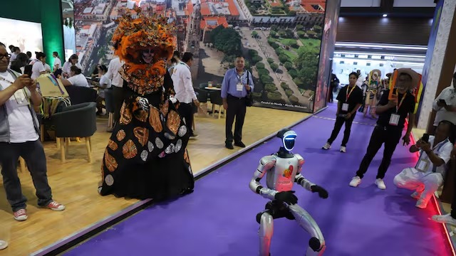 Un Robot Humanoide Plateado Avanza Sobre Una Alfombra Púrpura En El Tianguis Turístico México 2026. A Su Lado, Una Catrina Con Un Espectacular Tocado Naranja De Mariposas Monarca Observa La Interacción.