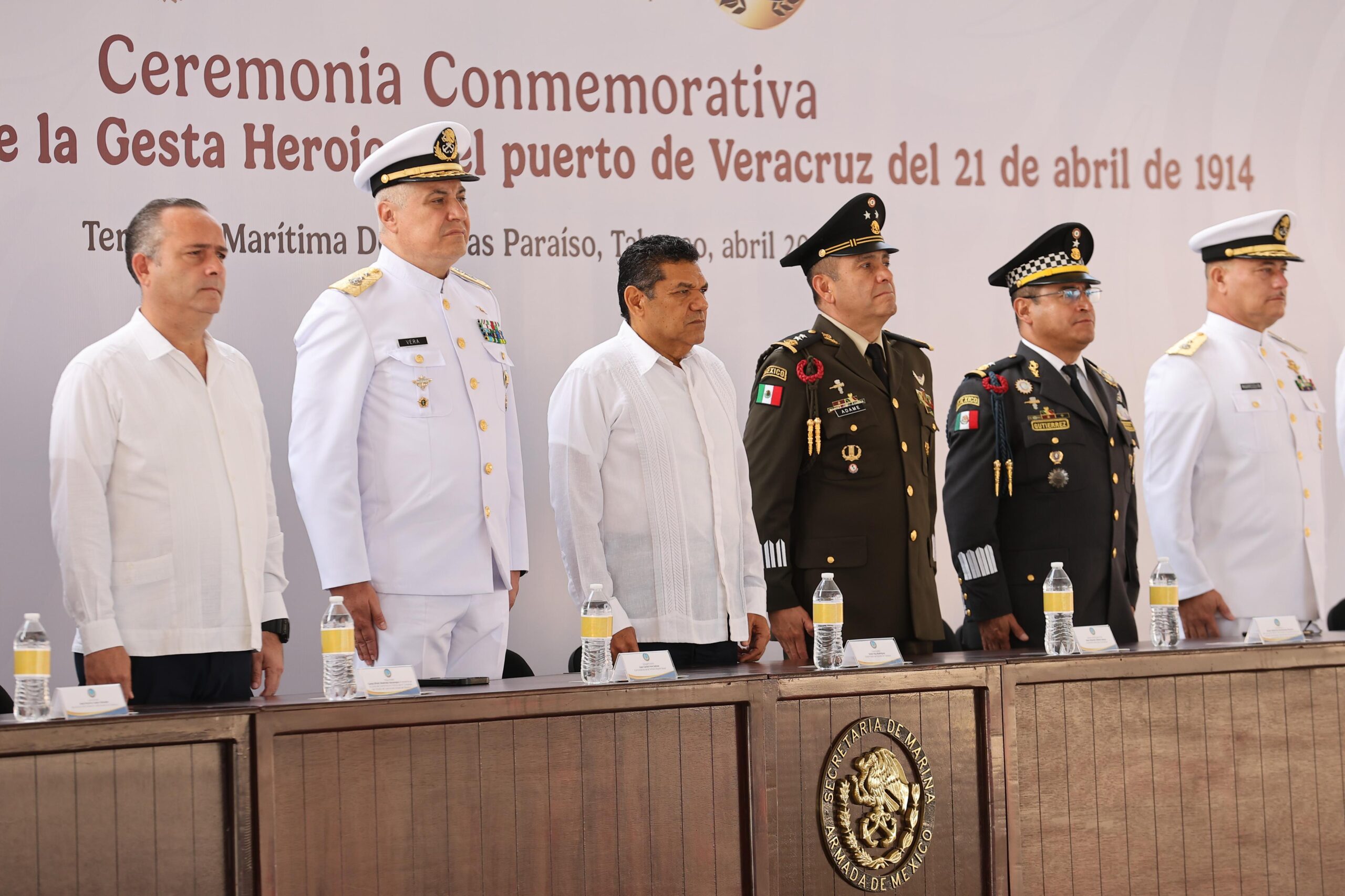 Autoridades Civiles Y Militares De Pie Frente A Un Presídium Durante La Ceremonia Conmemorativa De La Gesta Heroica Del Puerto De Veracruz. Al Centro Destaca El Gobernador De Tabasco, Javier May, Flanqueado Por Altos Mandos De La Secretaría De Marina Y La Secretaría De La Defensa Nacional En Uniformes De Gala. Al Fondo Se Observa Una Lona Con El Título Oficial Del Evento En Letras Guindas.