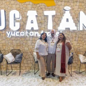 Tres Integrantes De La Delegación De Yucatán, Dos Mujeres Y Un Hombre, Posan Sonrientes Frente A Un Muro De Piedra Con El Nombre &Amp;Quot;Yucatán&Amp;Quot; En Letras Blancas Iluminadas Y El Logo &Amp;Quot;Yucatan.travel&Amp;Quot;.
