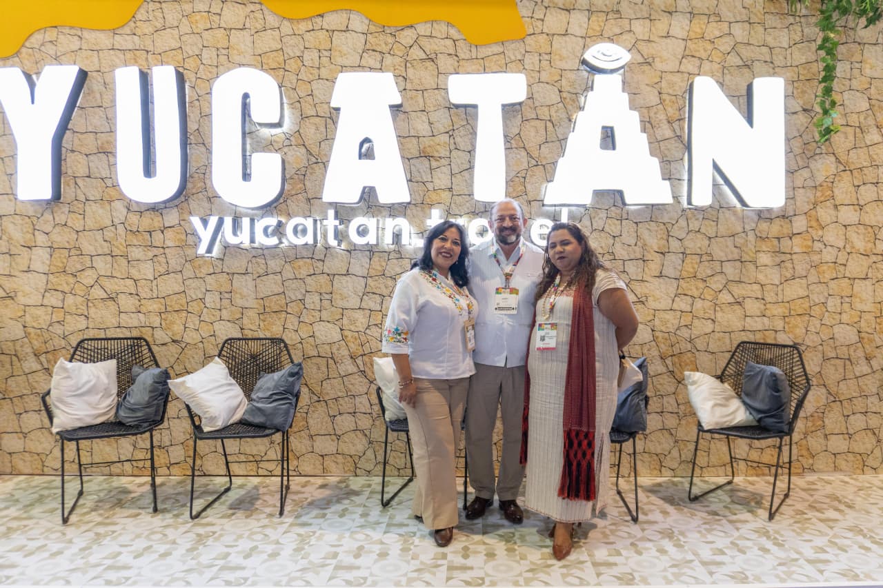 Tres Integrantes De La Delegación De Yucatán, Dos Mujeres Y Un Hombre, Posan Sonrientes Frente A Un Muro De Piedra Con El Nombre &Quot;Yucatán&Quot; En Letras Blancas Iluminadas Y El Logo &Quot;Yucatan.travel&Quot;.