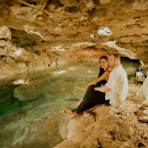 Yucatán hace historia: único estado en recibir doble galardón nacional a la innovación turística 2026 la revista del sureste mx Una Pareja Joven Posa Sentada A La Orilla De Un Cenote De Aguas Cristalinas Color Turquesa, Dentro De Una Caverna Iluminada Con Luz Cálida Que Resalta Las Formaciones De Estalactitas En El Techo. El Hombre Viste Una Guayabera Blanca Y Pantalón Oscuro, Mientras Que La Mujer Luce Un Vestido Negro De Noche. A La Derecha, Se Observa Una Mesa Preparada Para Una Cena Romántica De Lujo Con Mantel Blanco, Copas De Cristal Y Un Arreglo Floral, Creando Una Atmósfera Íntima Y Exclusiva En El Corazón Del Inframundo Maya En Yucatán.