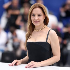 Fotografía En Plano Medio De La Actriz Mexicana Marina De Tavira Posando Sonriente Durante Una Sesión De Fotos En El Festival De Cannes. Viste Un Elegante Vestido Negro De Tirantes Delgados, Escote Recto Y Un Collar De Perlas Doradas De Varias Filas. Tiene El Cabello Castaño Claro Suelto Y Peinado De Lado. Sus Manos Descansan Sobre Una Barandilla Blanca En Primer Plano, Mientras Que El Fondo Muestra De Forma Desenfocada A Fotógrafos Y Asistentes Del Evento Sobre Un Fondo Azul Corporativo.