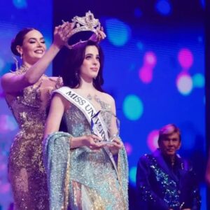 Fotografía Del Momento En Que Fátima Bosch, Miss Universo 2025, Coloca La Corona A Su Sucesora Sobre Un Escenario Iluminado. La Nueva Reina Viste Un Elegante Vestido De Gala Azul Claro Con Pedrería Y Porta La Banda De Miss Universe. Al Fondo Se Observa A Osmel Sousa Y Otra Finalista En Un Ambiente De Celebración Con Luces De Colores.