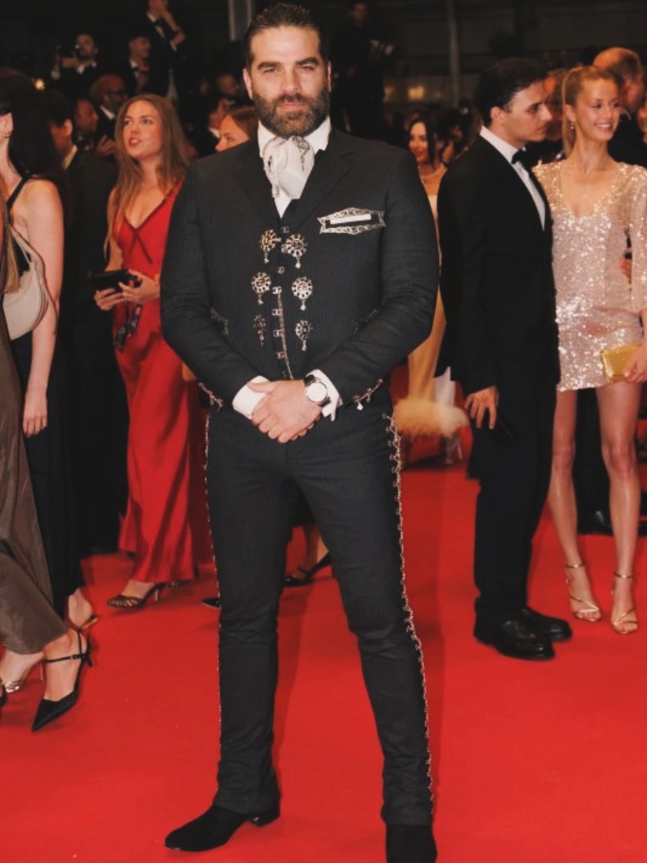 Alejandro nones impacta en el festival de cannes 2026 vestido con traje de charro la revista del sureste mx Fotografía De Plano Entero Del Actor Alejandro Nones Posando De Pie En El Centro De La Alfombra Roja Del Festival De Cannes. Viste Un Elegante Traje De Charro Mexicano De Gran Gala En Color Negro Con Sutiles Rayas Diplomáticas, Botonadura De Plata En El Chaleco Y Detalles Bordados Tradicionales A Los Costados Del Pantalón. Lleva Una Camisa Blanca Con Un Moño O Corbatón De Charro Claro Y Mantiene Las Manos Cruzadas Al Frente. Al Fondo Se Observa A Otros Asistentes Y Fotógrafos Desenfocados.