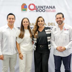 Una Fotografía Grupal En Interiores Que Muestra A Ocho Funcionarios De Pie Frente A Un Gran Telón De Fondo Blanco Durante Un Evento Oficial En Quintana Roo. En El Centro Destacan La Gobernadora Mara Lezama (Quinta Desde La Izquierda, Con Una Chaqueta De Rayas Blancas Y Negras Sobre Una Blusa Negra) Y La Presidenta Municipal Ana Paty Peralta (Tercera Desde La Izquierda, Vestida Totalmente De Blanco).