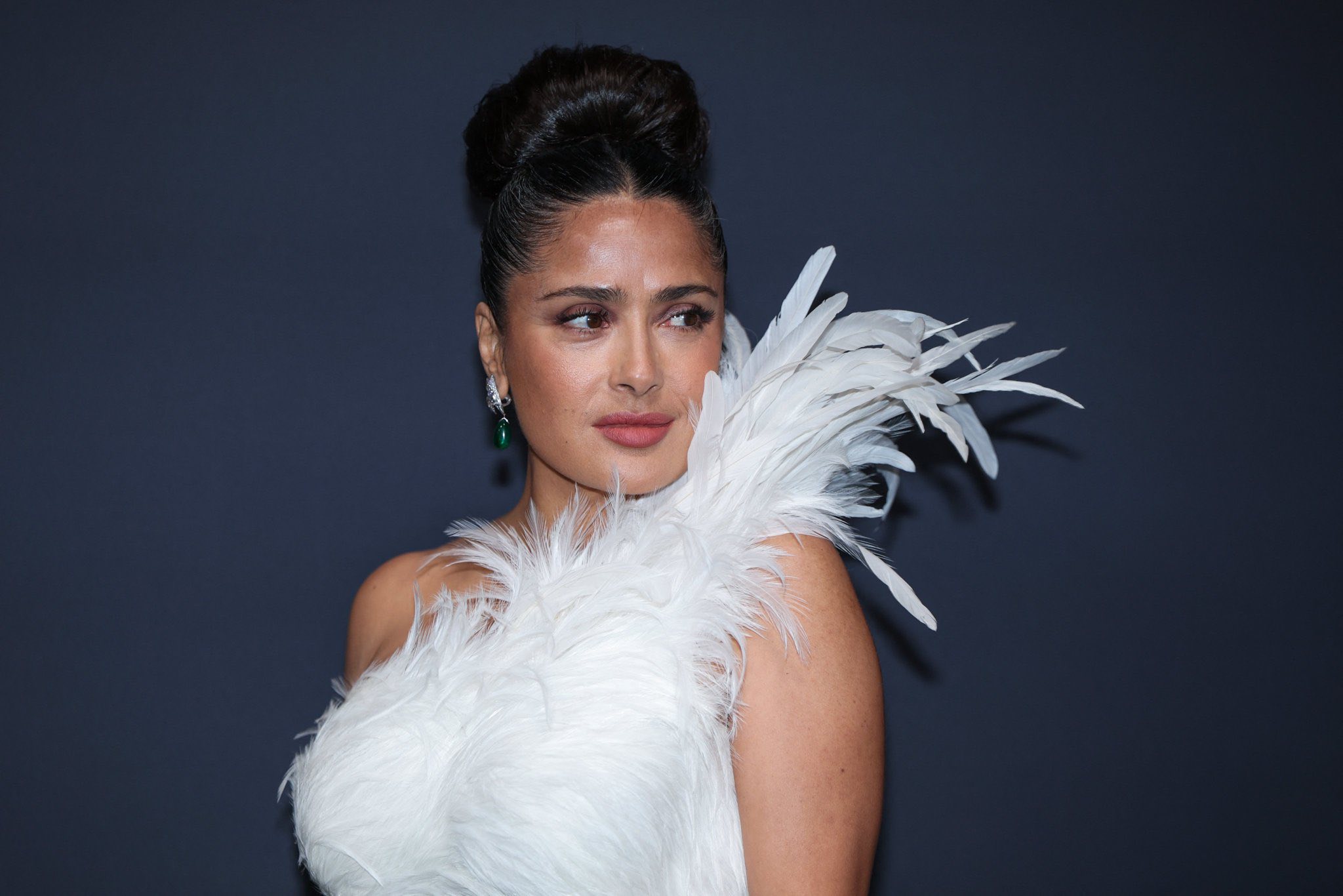 Retrato En Primer Plano Medio De La Actriz Mexicana Salma Hayek Posando De Tres Cuartos Frente A Un Fondo Liso De Color Gris Oscuro. La Intérprete Luce Un Peinado Recogido Alto Y Estilizado En Un Moño Voluminoso Y Pulido.