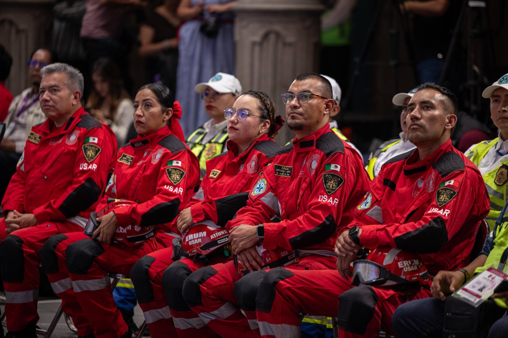 Primer Plano De Un Grupo De Elementos Del Escuadrón De Rescate Y Urgencias Médicas (Erum) Especializados En Búsqueda Y Rescate Urbano (Usar) Sentados Durante La Conferencia Del Primer Simulacro Nacional 2026. Los Rescatistas Visten Uniformes Tácticos De Color Rojo Brillante Con Insignias De La Policía Y Parches De &Quot;Usar&Quot;. En Sus Regazos Sostienen Cascos De Protección Rojos Con Visores Y Equipo De Seguridad. Al Fondo, Se Observa A Otros Servidores Públicos Con Chalecos De Alta Visibilidad, Todos Prestando Atención A La Presentación Oficial En Un Entorno Institucional.