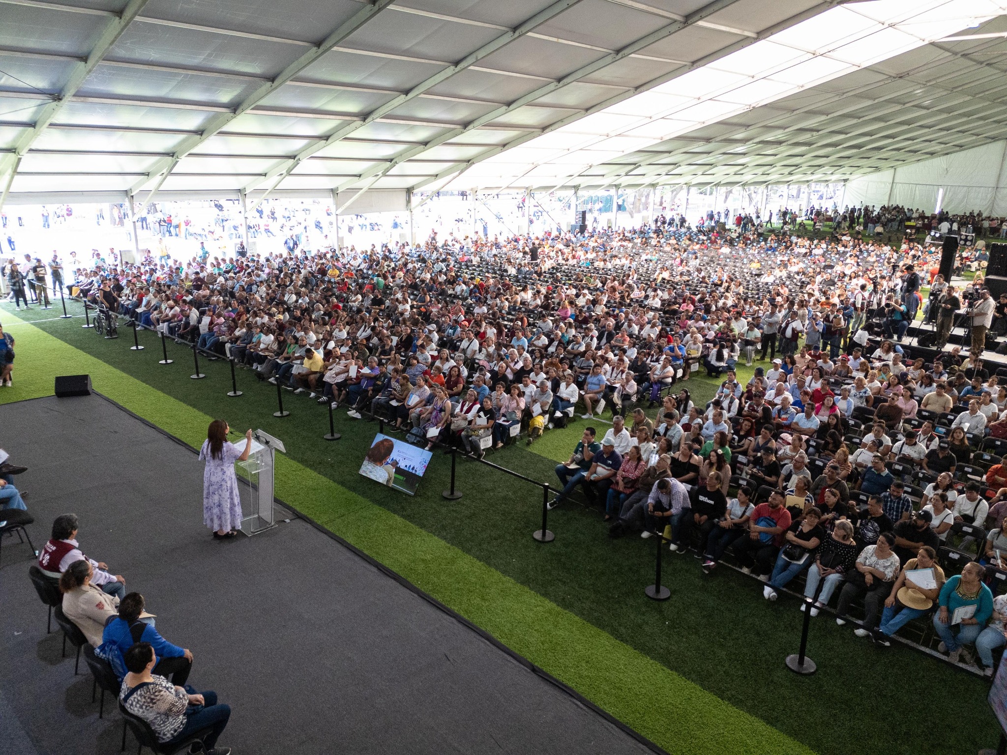 Una Toma Desde Un Ángulo Elevado Y Lateral Que Muestra Una Vista Panorámica De Un Evento Masivo Bajo Una Enorme Carpa Blanca Con Estructura Metálica. En El Lado Izquierdo, Sobre Un Escenario Elevado Con Piso Gris, La Jefa De Gobierno Clara Brugada Aparece De Espaldas, Hablando Desde Un Podio Transparente Hacia Una Multitud Inmensa. J