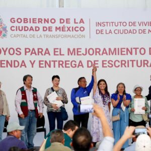 Una Toma De Plano Medio-Largo De Un Grupo De Ocho Personas De Pie En Un Escenario Frente A Un Gran Telón De Fondo Blanco Durante Un Evento Oficial Del Gobierno De La Ciudad De México. El Telón De Fondo Tiene Logotipos Y Texto Que Dice &Amp;Quot;Gobierno De La Ciudad De México&Amp;Quot;, &Amp;Quot;Capital De La Transformación&Amp;Quot;, &Amp;Quot;Instituto De Vivienda De La Ciudad De México&Amp;Quot; Y, En Letras Rojas Más Grandes, &Amp;Quot;Apoyos Para El Mejoramiento De Vivienda Y Entrega De Escrituras&Amp;Quot;.
