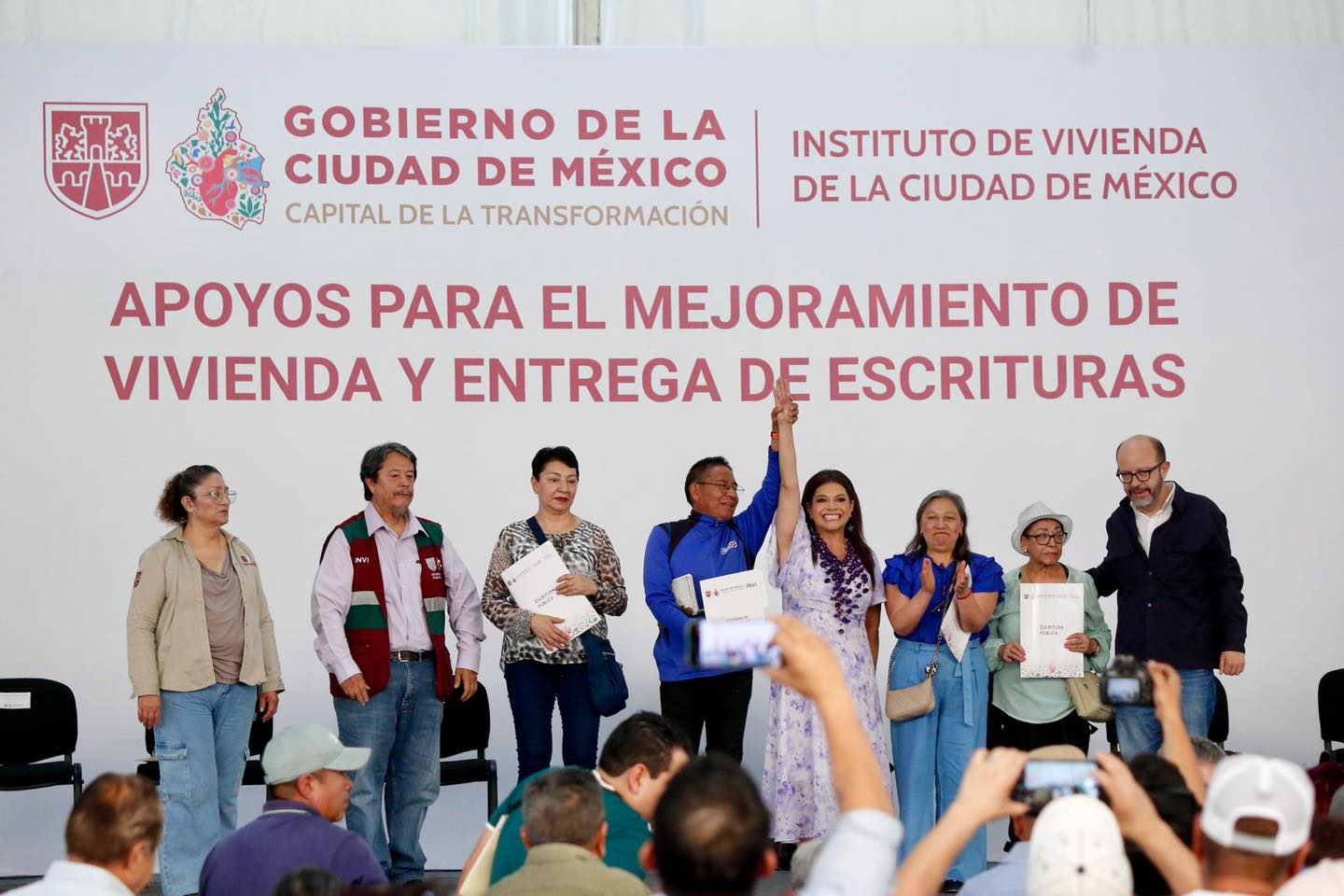 Una Toma De Plano Medio-Largo De Un Grupo De Ocho Personas De Pie En Un Escenario Frente A Un Gran Telón De Fondo Blanco Durante Un Evento Oficial Del Gobierno De La Ciudad De México. El Telón De Fondo Tiene Logotipos Y Texto Que Dice &Quot;Gobierno De La Ciudad De México&Quot;, &Quot;Capital De La Transformación&Quot;, &Quot;Instituto De Vivienda De La Ciudad De México&Quot; Y, En Letras Rojas Más Grandes, &Quot;Apoyos Para El Mejoramiento De Vivienda Y Entrega De Escrituras&Quot;.