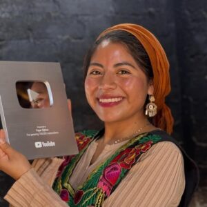 De santa maría tlahuitoltepec para el mundo: la oaxaqueña "ye ye ojitos" recibe el botón de plata de youtube la revista del sureste mx Un Primer Plano Vertical De Una Mujer Sonriente Con Cabello Castaño Oscuro Y Ojos Marrones, Que Lleva Un Pañuelo Naranja En La Cabeza Y Un Top Marrón Con Bordados Coloridos Alrededor Del Cuello. Sostiene Con Orgullo Un Premio De Youtube De Plata Que Dice &Amp;Quot;Presented To Yeye Ojitos For Passing 100,000 Subscribers&Amp;Quot; En Su Mano Izquierda. El Fondo Es Una Pared De Ladrillo Gris Oscuro Con Algunas Texturas Y Variaciones En La Iluminación.