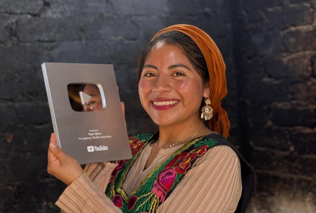 De santa maría tlahuitoltepec para el mundo: la oaxaqueña "ye ye ojitos" recibe el botón de plata de youtube la revista del sureste mx Un Primer Plano Vertical De Una Mujer Sonriente Con Cabello Castaño Oscuro Y Ojos Marrones, Que Lleva Un Pañuelo Naranja En La Cabeza Y Un Top Marrón Con Bordados Coloridos Alrededor Del Cuello. Sostiene Con Orgullo Un Premio De Youtube De Plata Que Dice &Quot;Presented To Yeye Ojitos For Passing 100,000 Subscribers&Quot; En Su Mano Izquierda. El Fondo Es Una Pared De Ladrillo Gris Oscuro Con Algunas Texturas Y Variaciones En La Iluminación.