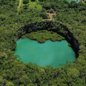 Fotografía Aérea En Plano General Del Cenote El Zacatón. Se Observa Una Colosal Fosa Circular De Aguas Color Verde Turquesa, Rodeada Por Paredes De Roca Escarpada Y Una Densa E Interminable Selva Verde Bajo Un Cielo Parcialmente Nublado. En La Superficie Del Agua Flotan Pequeñas Islas Verdes De Zacate.