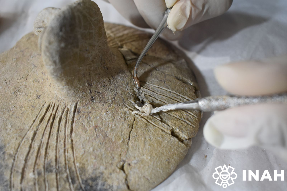 Descubren urna maya con figura de búho que revela rituales funerarios en la península de yucatán la revista del sureste mx Se Observa Un Primer Plano Detallado De La Tapa De La Urna Arqueológica De Arcilla Con Líneas Incisas Decorativas. Manos Con Guantes Quirúrgicos De Látex Blanco Utilizan Instrumentos Metálicos De Precisión Fina, Similares A Espátulas Y Ganchos De Restauración, Para Limpiar Con Sumo Cuidado Los Sedimentos Acumulados Y Consolidar Las Grietas De La Pieza Prehispánica. En La Esquina Inferior Derecha Se Incluye El Logotipo Institucional Blanco Del Inah.