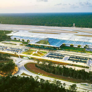 Una Vista Aérea Del Aeropuerto Internacional De Tulum &Amp;Quot;Felipe Carrillo Puerto&Amp;Quot;, Un Edificio Terminal Moderno Y Amplio En El Centro, Con Un Techo Distintivo De Color Azul Y Blanco Con Motivos Inspirados En El Diseño Maya. La Terminal Está Iluminada En La Oscuridad, Rodeada Por Caminos De Acceso Curvos, Estacionamientos Llenos De Vehículos Y Áreas Ajardinadas. Detrás De La Terminal Se Extiende La Pista De Aterrizaje Y Las Calles De Rodaje, Y Al Fondo, Un Denso Bosque Tropical Bajo Un Cielo Crepuscular