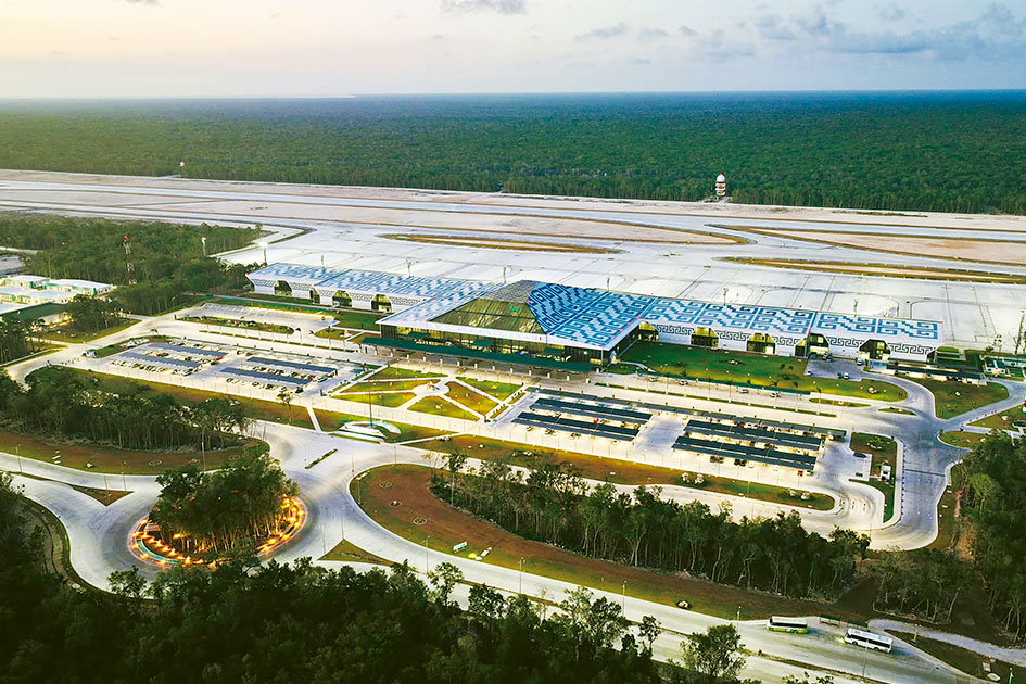 Una Vista Aérea Del Aeropuerto Internacional De Tulum &Quot;Felipe Carrillo Puerto&Quot;, Un Edificio Terminal Moderno Y Amplio En El Centro, Con Un Techo Distintivo De Color Azul Y Blanco Con Motivos Inspirados En El Diseño Maya. La Terminal Está Iluminada En La Oscuridad, Rodeada Por Caminos De Acceso Curvos, Estacionamientos Llenos De Vehículos Y Áreas Ajardinadas. Detrás De La Terminal Se Extiende La Pista De Aterrizaje Y Las Calles De Rodaje, Y Al Fondo, Un Denso Bosque Tropical Bajo Un Cielo Crepuscular