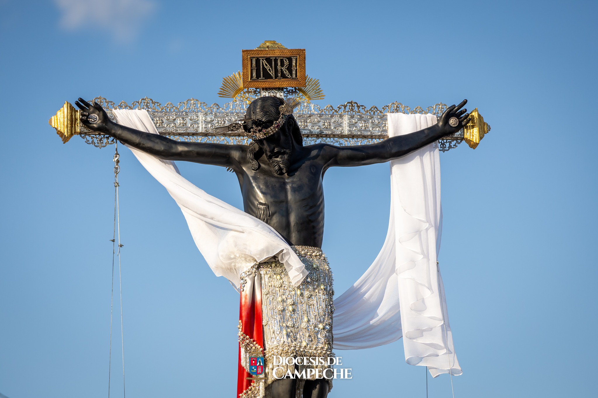 Una Toma De Plano Medio Del Cristo Negro De San Román, Una Escultura Religiosa De Ébano O Madera Oscura, Crucificada En Una Cruz Adornada Con Intrincados Detalles De Plata. La Figura De Cristo Viste Un Perizoma (Paño De Pureza) Blanco Bordado Con Pedrería Y Perlas, Y Tiene Una Tela Blanca Larga Y Ligera Que Cuelga Elegantemente De Sus Brazos Extendidos.