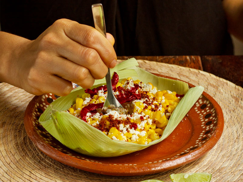 Una Mano Sostiene Una Cuchara Para Comer Un Esquite Servido Dentro De Una Hoja De Maíz (Totomoxtle). El Platillo, Colocado Sobre Un Plato De Barro Artesanal, Contiene Granos De Elote Amarillo, Queso Desmoronado, Chile En Polvo Y Chiles Secos Enteros. Todo Reposa Sobre Un Mantel De Palma Tejido.