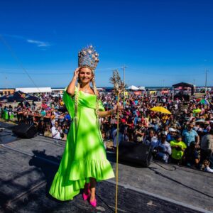 En El Centro, Una Mujer Joven Posa Sonriente Vistiendo Un Llamativo Vestido Largo Color Verde Neón Oaxaqueño Y Tacones Rosa Encendido. Porta Una Gran Corona Dorada Ornamentada Con Motivos Marinos Y Sostiene Un Cetro Dorado En Su Mano Izquierda.